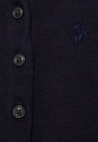 Marinbl�å stickad kofta med fem svarta knappar, texturerad tyg och en subtil broderad logotyp. Slät struktur, avslappnad design.