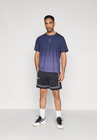 Μπλε μπλουζάκι με διαβάθμιση και κοντά μανίκια, συνδυασμένο με μαύρο σορτς από δίχτυ με λευκή επένδυση, και μαύρα και άσπρα αθλητικά παπούτσια.