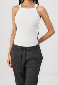 Bílý žebrovaný top na ramínka s širokými popruhy ve spojení s tmavě šedými tepláky na šňůrku, které mají kapsy a volný střih.