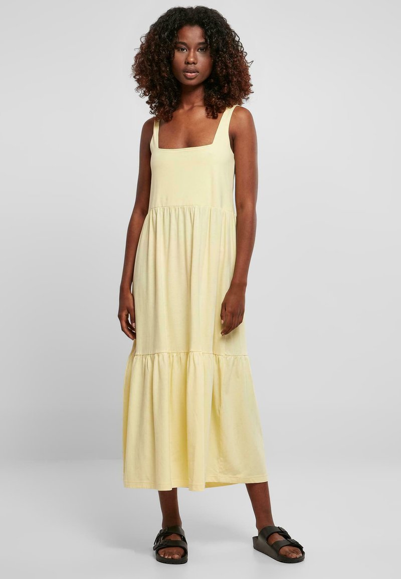 Mujer con cabello rizado vestida con un vestido midi sin mangas de color amarillo pálido y sandalias negras sin tacón, sobre un fondo liso gris claro.