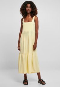 Geel katoen maxi-jurk met een vierkante halslijn, mouwloos ontwerp, gathered taille en gelaagde onderzoom, gecombineerd met zwarte sandalen.
