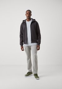 Felpa con zip grigia scura sopra una t-shirt grigia chiara, abbinata a pantaloni grigi chiari e scarpe sportive verdi. Design semplice, texture liscia.
