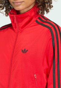 Röd atletisk jacka med hög krage, med svarta sidostripes och ficka med dragkedja. Den har en broderad logotyp på vänster bröst.