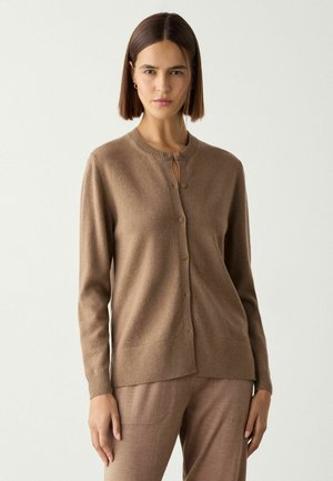Frau mit glattem dunklem Haar trägt einen geknöpften braunen Cardigan und passende Hose, steht vor einem schlichten hellen Hintergrund.