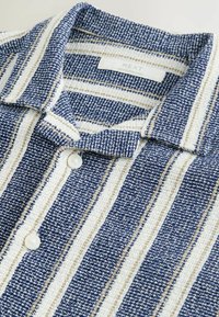 Chemise rayée bleu marine et blanche en tissu texturé, dotée d'un col et d'une patte de boutonnage. Comprend des rayures beige discrètes.