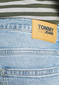 Světle modré džínové kalhoty s hnědou koženou nášivkou s nápisem "TOMMY JEANS" v černé barvě. Pás je částečně zakrytý pruhovanou zeleno-šedou košilí.