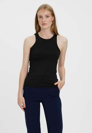 Blonde vrouw in een mouwloos zwart topje en donkerblauwe broek, staand met één hand in haar zak tegen een effen lichte achtergrond.