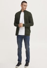 Grøn ribbet lynlåsjakke over en hvid t-shirt, sat sammen med blå jeans og sorte sneakers. Glat tekstur med en afslappet pasform.