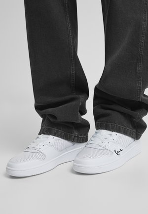 Weiße perforierte Sneaker mit flachen Sohlen, einem gestickten Logo und einem Obermaterial aus Stoff, kombiniert mit locker sitzenden schwarzen Jeanshosen.