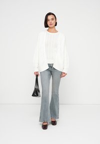 Λευκό oversized ζακέτα με πλεκτό καλώδιο πάνω από λευκό ριμπ τοπ, συνδυασμένο με ανοιχτό μπλε καμπάνες τζιν και σκούρα καφέ λουστρίνια. Συμπεριλαμβάνεται μαύρη τσάντα χειρός.