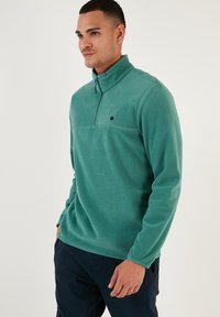 Pullover in pile verde con colletto a zip quart, caratterizzato da un logo nero, una texture morbida e maniche lunghe. Abbinato a pantaloni blu navy.