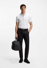 Witte polo met een ritssluiting bij de hals en lichte bies, gecombineerd met zwarte broek en een stijlvolle zwarte duffeltas. Model draagt zwarte sneakers.