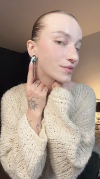 Strieborné náušnice s oblým dizajnom a tyrkysovým akcentom. Modelka má na sebe krémový pletený sveter a ukazuje si tetovania na prstoch.