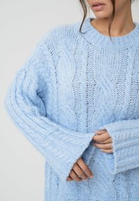 Ljusblå stickad tröja med ett kabelstickat mönster, vida ärmar och ribbad halsringning. Mjuk textur med en lös passform.