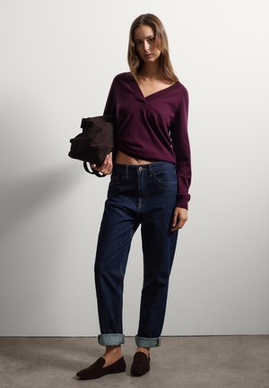 Donna con un maglione a V color prugna, jeans blu scuri risvoltati, mocassini marroni, che tiene una borsa in camoscio marrone contro uno sfondo bianco.