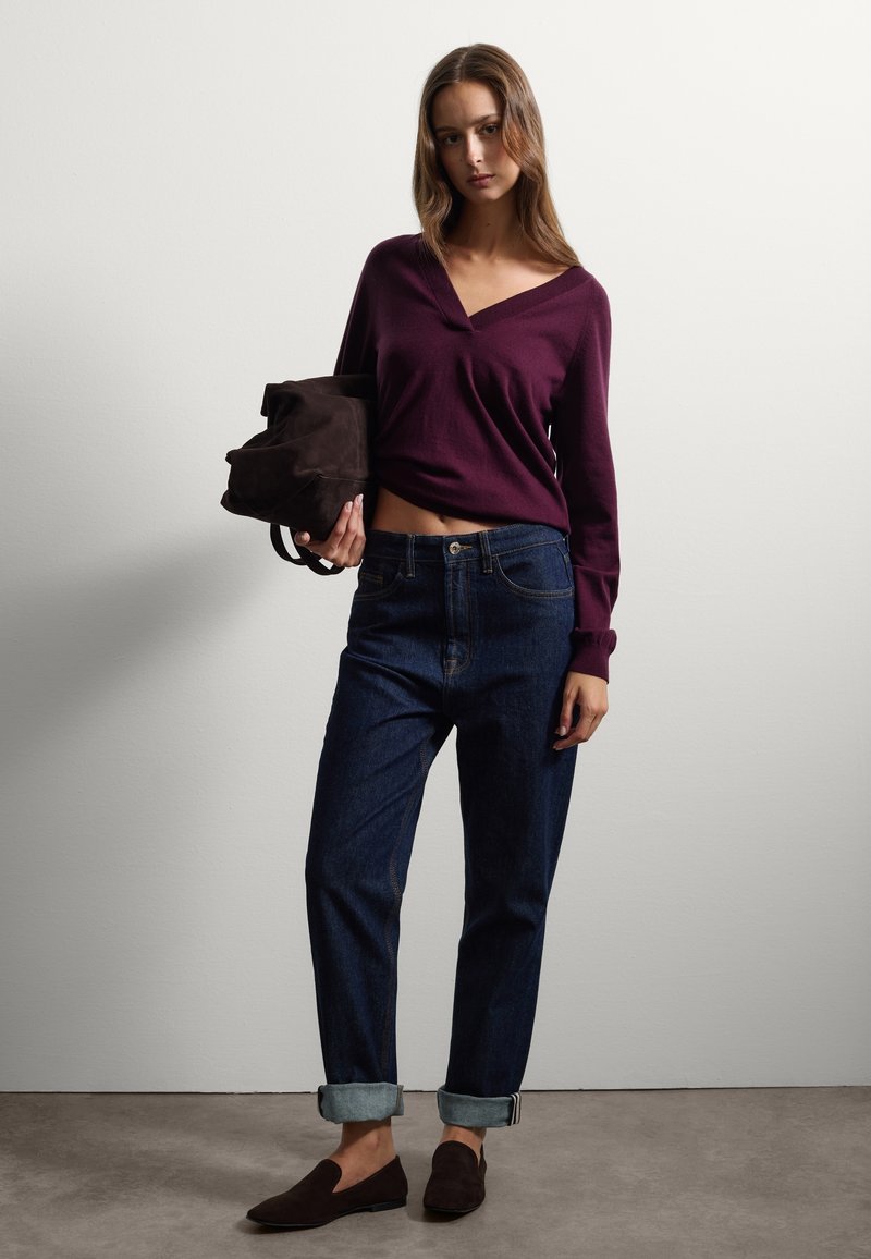 Femme portant un pull violet à col en V, un jean bleu foncé retroussé, des mocassins marron, tenant un sac en daim marron contre un mur blanc.
