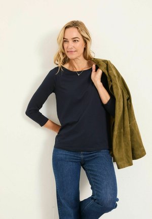 Navy-blaues Langarmoberteil mit einem Bootsausschnitt, kombiniert mit einer strukturierten olivgrünen Wildlederjacke und blauen Denim-Jeans. Einfaches Design.