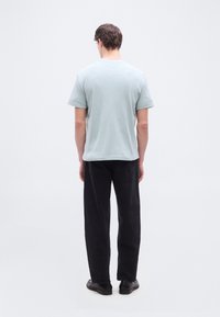 Άνδρας που στέκεται γυρισμένος με την πλάτη προς τα εμπρός, φορώντας ανοιχτό μπλε πουκάμισο με κοντά μανίκια, μαύρο παντελόνι και μαύρα παπούτσια, μπροστά σε ένα απλό λευκό φόντο.