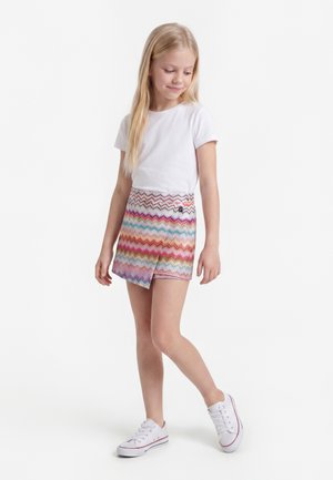 Fille blonde debout, portant un t-shirt blanc, une jupe colorée à motif zigzag et des baskets blanches, regardant vers le bas avec un léger sourire.