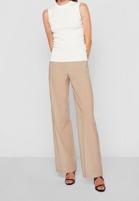 Mujer vestida con una blusa sin mangas blanca de tela acanalada, pantalones beige de pierna ancha y tacones negros abiertos, de pie frente a un fondo claro y liso.