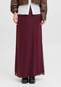 Falda maxi burdeos en tejido ligero y fluido. Presenta un dobladillo en forma de ondas y se combina con una chaqueta de cuero marrón y botas negras.