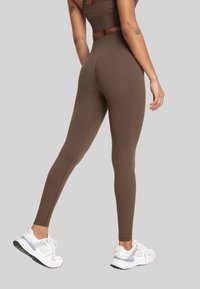 Kobieta w brązowych legginsach o wysokim stanie i dopasowanej bluzce, stojąca z jedną nogą lekko zgiętą, w białych trampkach, z widocznymi tatuażami na ramionach.