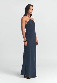 Halskleid in Marineblau mit figurbetonter Silhouette und Knöchellänge. Glatte Textur mit minimalen Details, getragen mit schwarzen und goldenen Accessoires.