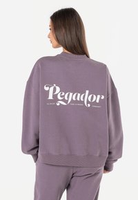 Overdimensjonert lilla hettegenser med hvit tekst på ryggen som sier "Pegador" med ekstra tekst. Mykt stoff, ribbede mansjetter og kant.