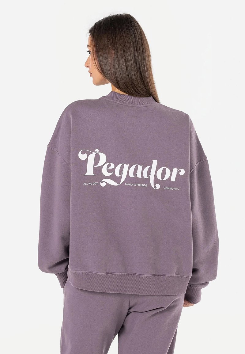 Overdimensjonert lilla hettegenser med hvit tekst på ryggen som sier "Pegador" med ekstra tekst. Mykt stoff, ribbede mansjetter og kant.