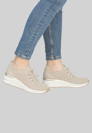 Person, die beige gestrickte Sneakers mit weißen Sohlen und hellblauen Skinny-Jeans vor einem grauen Hintergrund trägt.