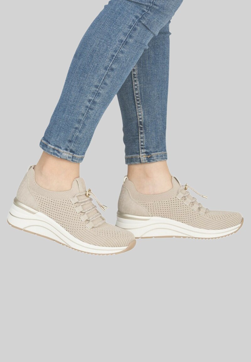 Personne portant des baskets en tricot beige avec des semelles blanches et un jean skinny bleu clair sur un fond gris.