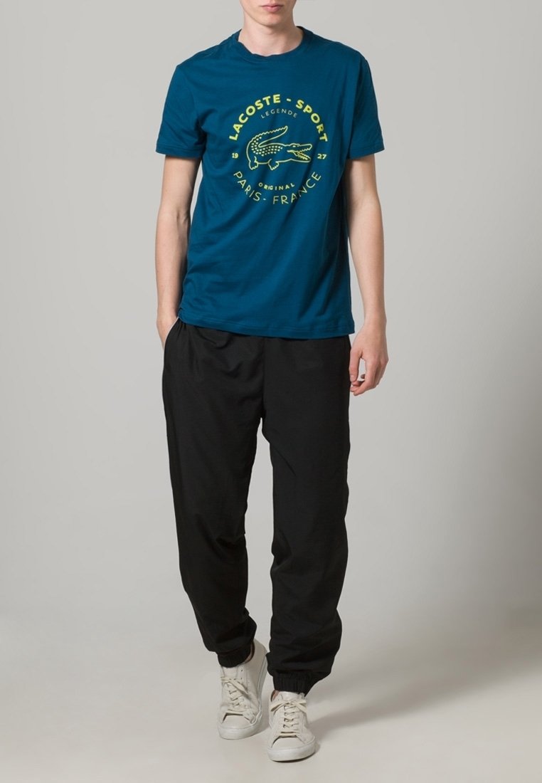 Jeune homme portant un t-shirt bleu Lacoste Sport avec logo, un pantalon de jogging noir et des baskets blanches, debout devant un fond gris uni.
