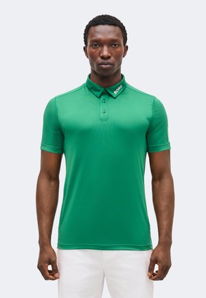 Άνδρας που φοράει πράσινο πόλο με κοντά μανίκια και το κείμενο «BORG» στον γιακά, καθώς και λευκό παντελόνι, στέκεται μπροστά σε μονόχρωμο φόντο.