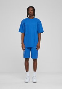 Blå oversized t-skjorte og matchende shorts laget av myk bomull, med en avslappet passform. Brukt med hvite sneakers og sokker.