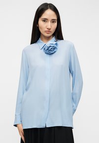 Ανοιχτό μπλε μπλούζα με μακριά μανίκια από διάφανο ύφασμα, με μια δομημένη εφαρμογή ρόδου στο αριστερό στήθος, που διαθέτει κολάρο με κουμπιά.