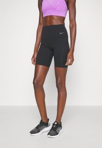 Shorts de sport noirs avec une taille haute, associés à un crop top géométrique violet et des chaussures de course noires avec des semelles blanches.