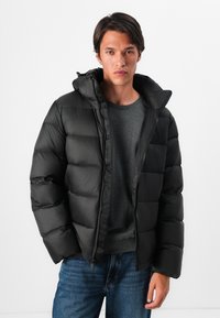 Man in een zwarte pufferjack over een donkergrijze trui en blauwe jeans, staand tegen een effen witte achtergrond.