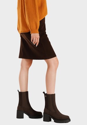 Donna che indossa una gonna marrone al ginocchio, un maglione arancione e stivaletti neri con tacco, in piedi su uno sfondo semplice.