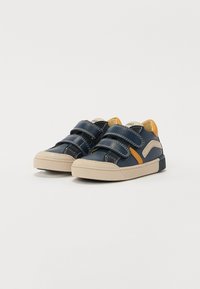 Adidași din piele bleumarin cu două curele Velcro, accente bej și o talpă din cauciuc texturată. Prezintă cusături contrastante și un tab pentru călcâi portocaliu.