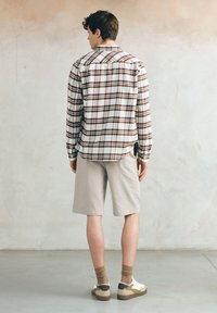 Mężczyzna widziany z tyłu, ubrany w białą i brązową koszulę w kraty, beżowe szorty do kolan, brązowe skarpetki i białe trampki, na tle gładkiej ściany.