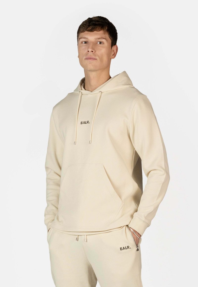 Beige hættebluse lavet af blødt stof, med en forlommelomme og snore. Brandnavnet "BALR." er broderet på brystet.