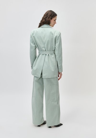 Γυναίκα με μακριά σκούρα μαλλιά, ντυμένη με ένα ανοιχτό πράσινο σακάκι με ζώνη και παντελόνια με φαρδιά μπατζάκια, όρθια και στραμμένη με την πλάτη προς τα πίσω, πάνω σε απλό φόντο.