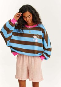 Světle modrý a hnědý pruhovaný svetr s růžovými manžetami a designem mušle. V kombinaci se světle růžovými leninovými šortkami, které jsou ohrnuté na spodním okraji.