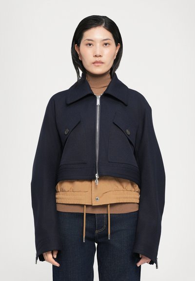 Veste courte en tissu bleu marine foncé avec des poches poitrine distinctes, fermeture éclair argentée et ourlet en couches beige. Associée à un pull col roulé marron et un jean.