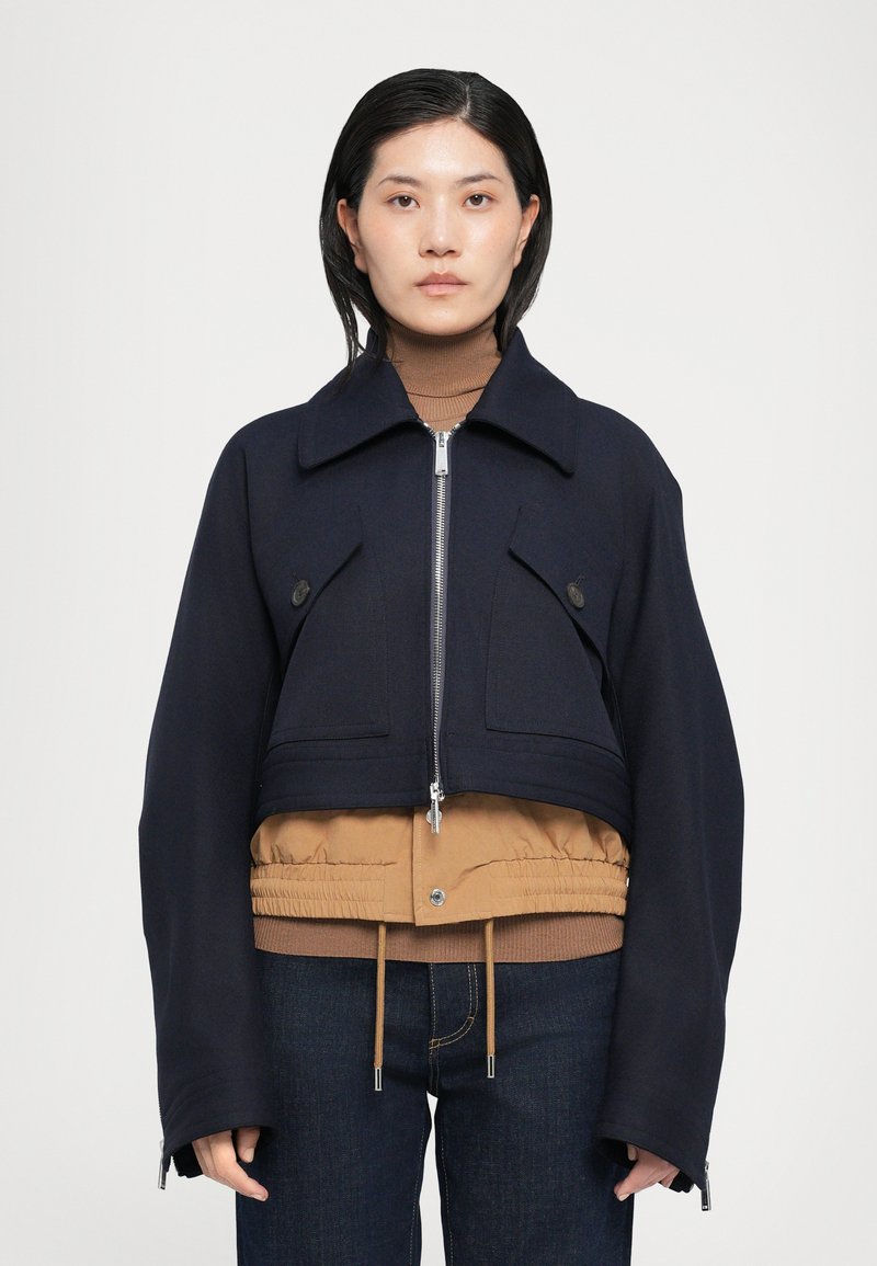Chaqueta recortada en tejido azul marino oscuro con bolsillos en el pecho distintivos, cremallera plateada y dobladillo en capas color marrón. Combinada con un cuello de tortuga marrón y denim.