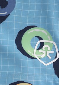 Blått tyg har cirkulära donutmönster i grönt och mörkblått, med en vit sköldlogotyp som innehåller en personikon nära botten.