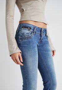 Jean en denim bleu avec une coupe slim, dotée d'une poche zippée, de surpiqûres contrastées et d'une taille haute. Texture lisse et délavage.