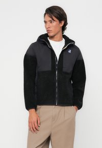 Veste noire avec des manches en peluche texturées et une capuche, présentant un panneau avant lisse. Fermeture éclair, détail de logo sur la poitrine, associée à un pantalon beige.