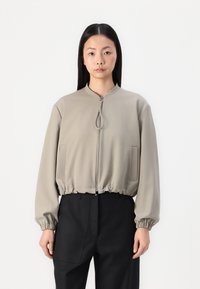 Giacca bomber zip con un leggero beige, con orlo e polsini elasticizzati, dotata di due tasche laterali e colletto rialzato. Tessuto dalla superficie morbida.