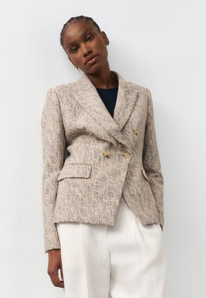 Žena s pletenými vlasmi nosí béžové textúrované dvojradové sako cez čierny top a biele nohavice s vysokým pásom, stojaca pri bielej stene.